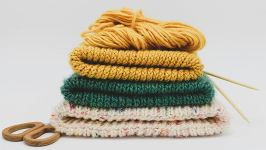 A pile of knitting projects, knitting needles and some wooden scissors.