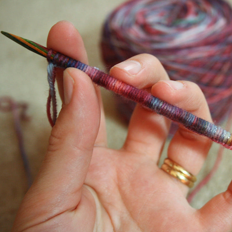 Hand holding a knitting needle with yarn, blurred yarn ball in the background