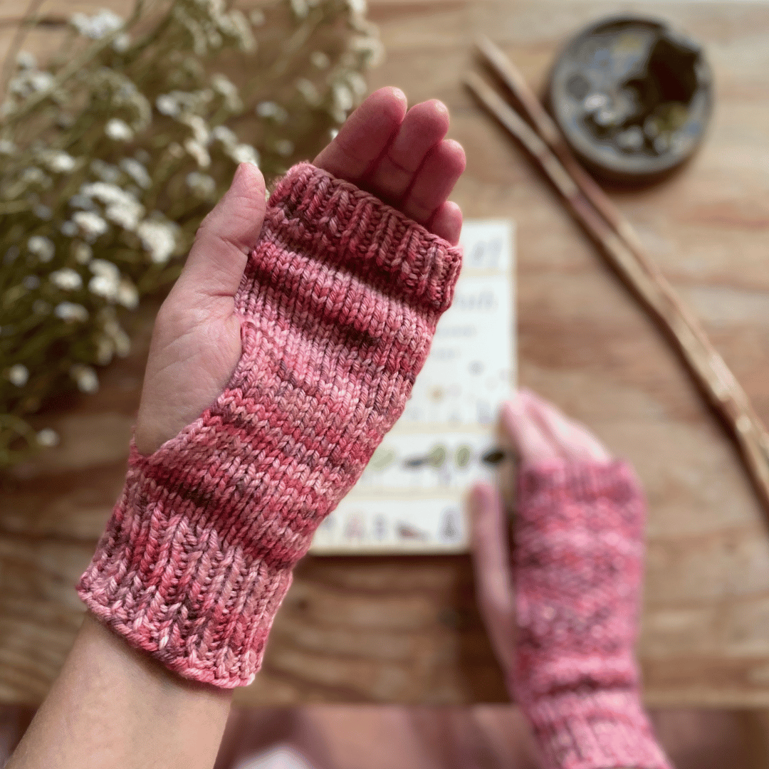 Two hands wearing hand dyed, knitted fingerless mitts.
