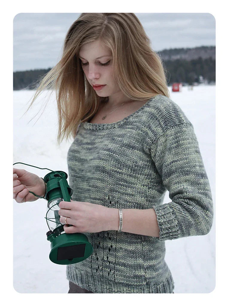 Young woman wearing a hand-dyed grey-green knit sweater holding a green lantern outdoors in snowy landscape