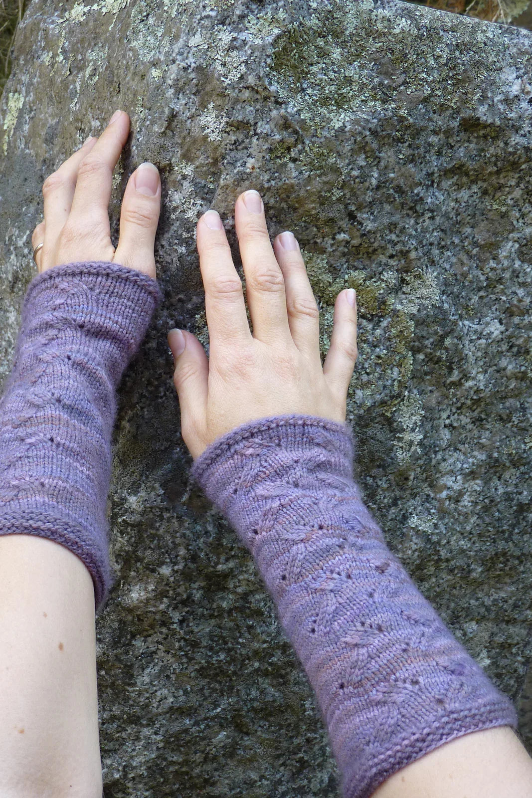 Hands wearing hand-dyed purple knitted wrist warmers resting on a textured mossy rock