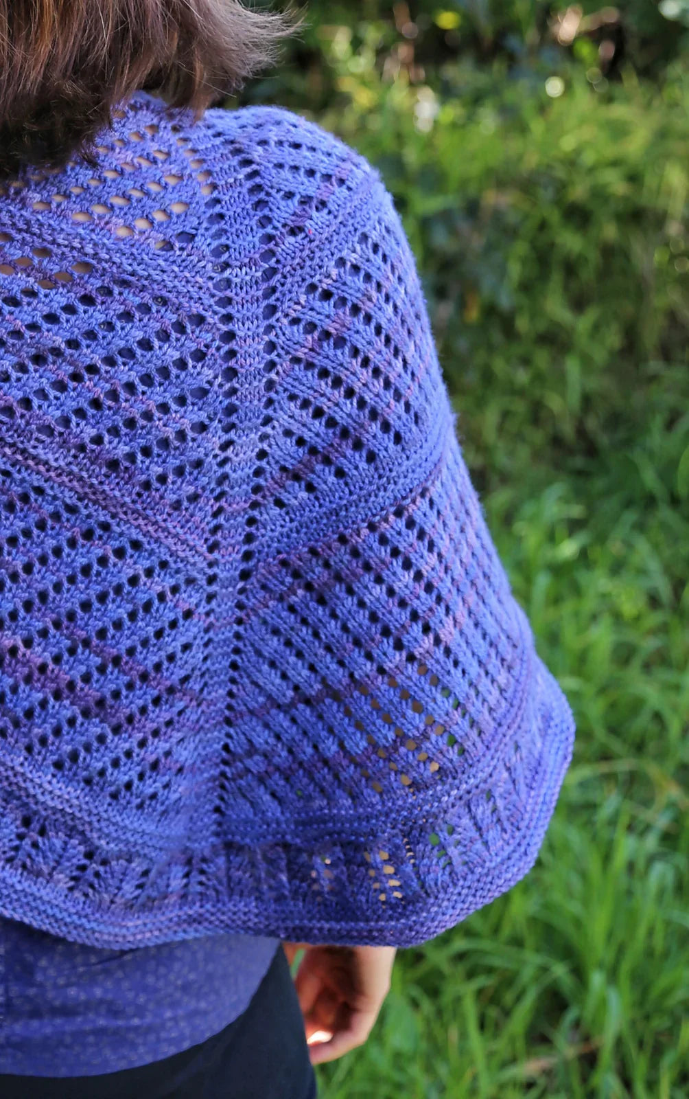 Close-up of a person wearing a hand-dyed blue lace-patterned knitted shawl outdoors with green foliage background