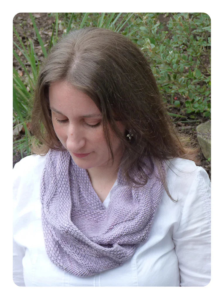 Woman wearing hand-dyed lavender knitted cowl scarf outdoors with greenery background