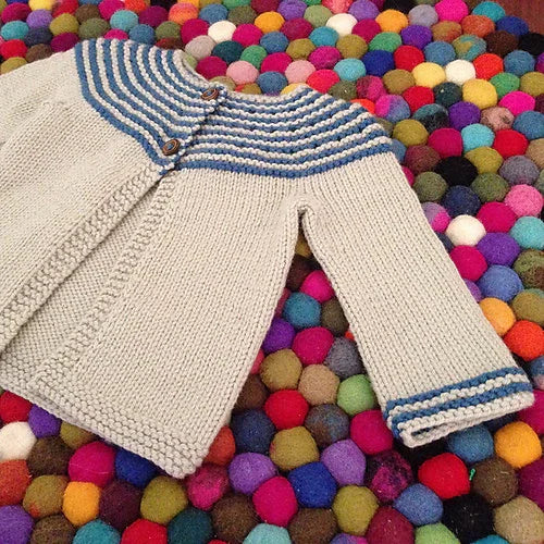 Hand-knitted cream baby cardigan with blue striped yoke and cuffs, displayed on colorful felt balls rug, made with hand-dyed yarn