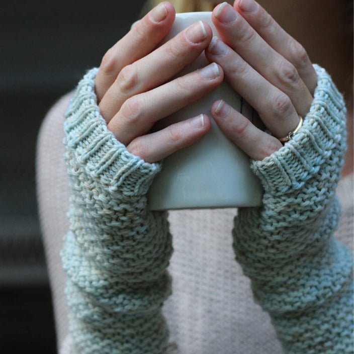 Two hands wearing hand knitted fingeless mittens holding a mug.