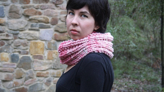 Woman wearing a hand-dyed pink chunky knit scarf outdoors near a stone wall and greenery