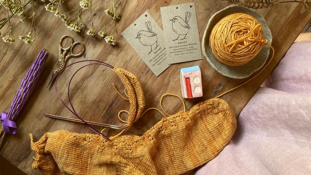 A hand knitted sock and some knitting on a wooden table with two augustbird yarn tags with illustrations of birds on them.