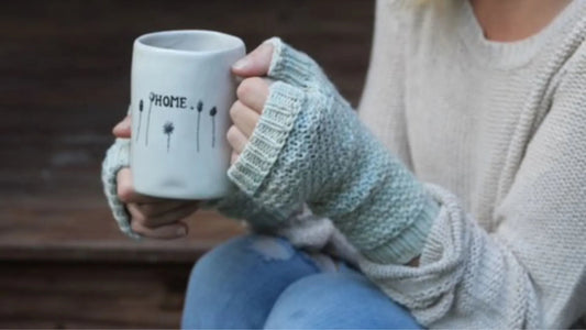 Person wearing hand-dyed fingerless gloves holding white mug labeled HOME in cozy knit sweater