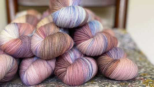 Soft hand-dyed yarn skeins in muted pink, purple, blue, and beige tones piled on floral fabric