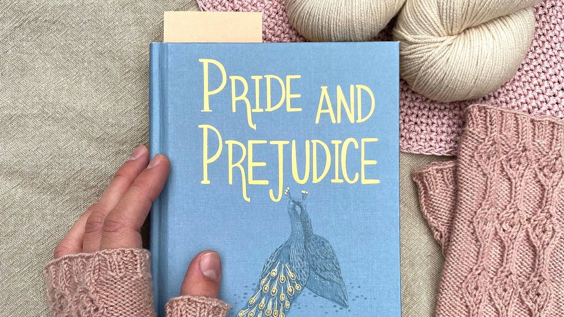 Hand holding a blue book titled Pride and Prejudice with hand-dyed yarn and pink knitted items on linen surface