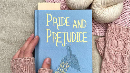 Hand holding a blue book titled Pride and Prejudice with hand-dyed yarn and pink knitted items on linen surface