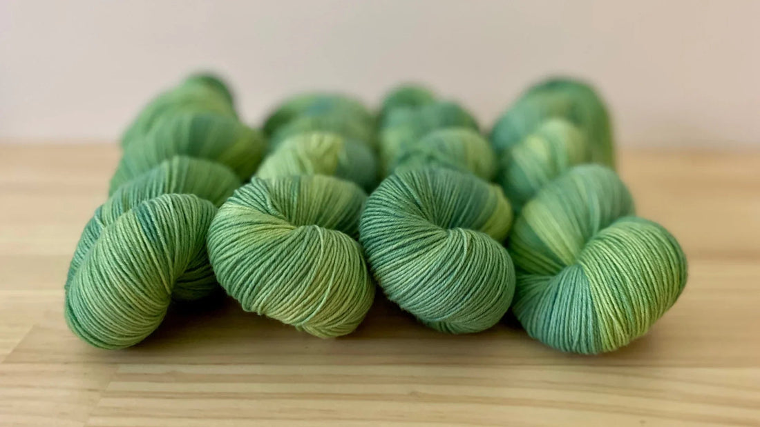 Hand-dyed green yarn skeins arranged on light wood surface, showcasing soft, variegated colorways