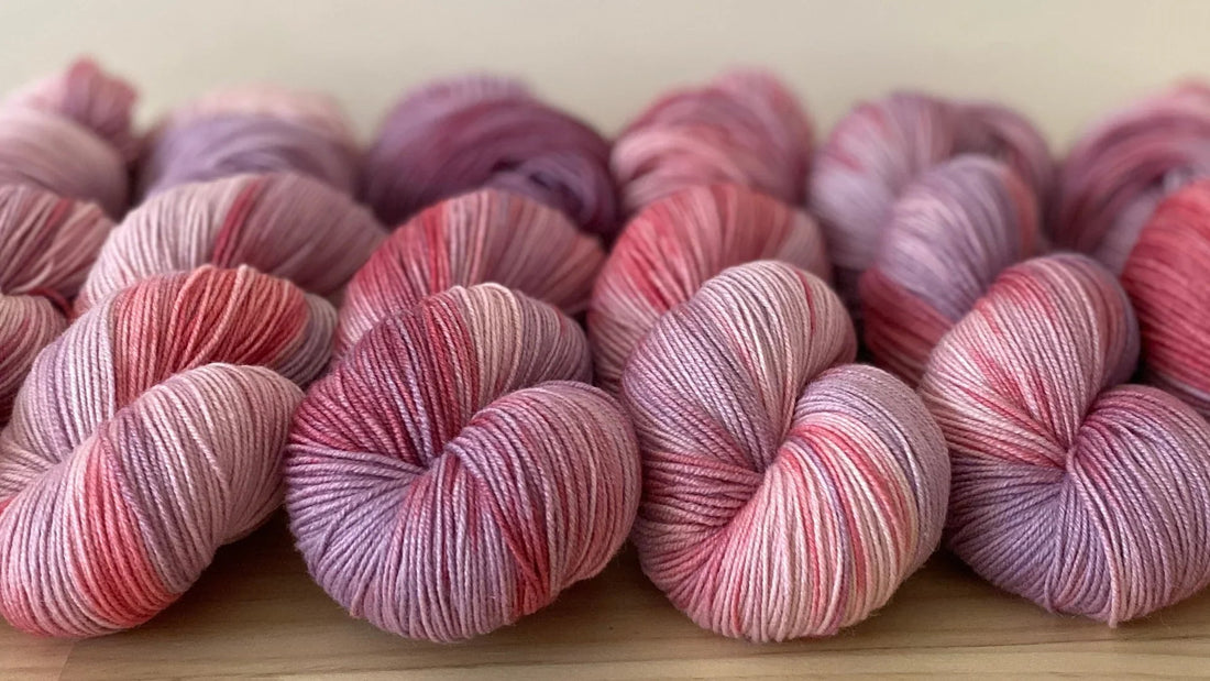 Hand-dyed yarn skeins in soft pink and purple tones arranged on a wooden surface