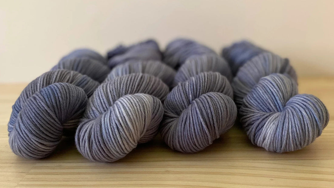 Multiple skeins of hand-dyed soft yarn in varying shades of blue and gray on a wooden surface
