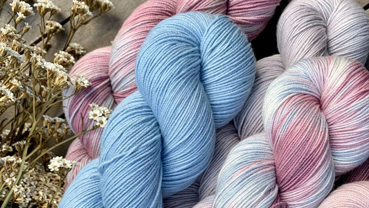 Hand-dyed yarn skeins in soft blue, pink, and white hues with dried flowers on wooden surface