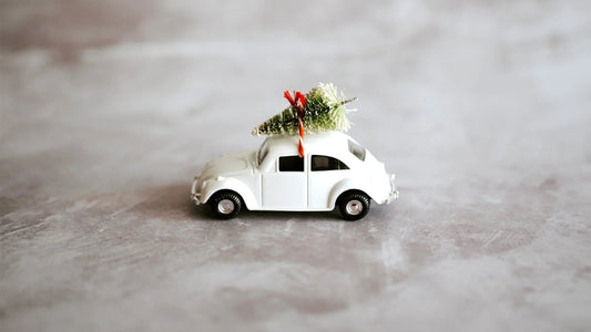 White toy car carrying a small green Christmas tree tied with red hand-dyed yarn on gray surface