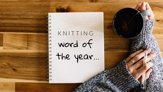 Notebook on wooden table with text knitting word of the year, hand in gray knitted sweater holding pen and cup of tea