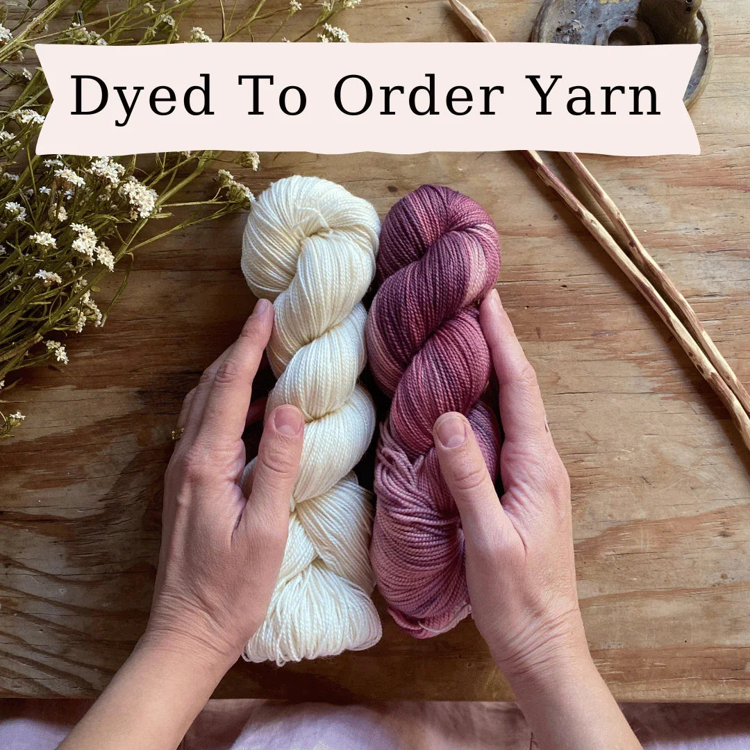 Hands holding two skeins of hand-dyed yarn in cream and purple on a wooden table with dried flowers