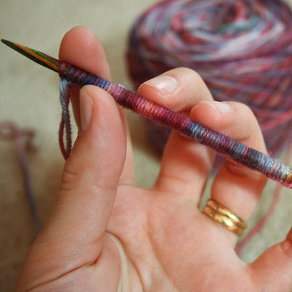 Hand holding a knitting needle with yarn, blurred yarn ball in the background