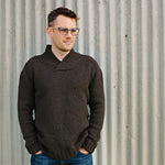 Man wearing a brown knitted jumper standing infant of a corrugated iron fence.