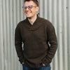 Man wearing a brown knitted jumper standing infant of a corrugated iron fence.